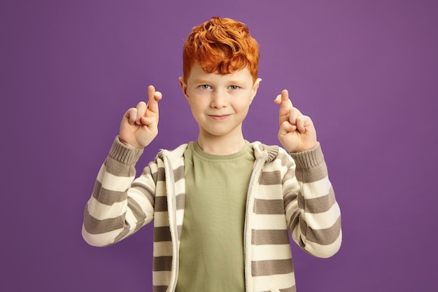 Handsome Little Boy has Red Hair Crossing his Fingers Making Wish Standing on Violet Isolated