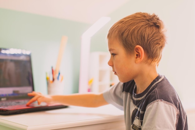 the handsome little boy is sitting at his desk at home and using a laptop for online learning