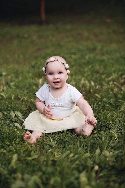 Handsome little girl with short fair hair and pretty smile in white dress sits on a grass in the park in summer and smiles