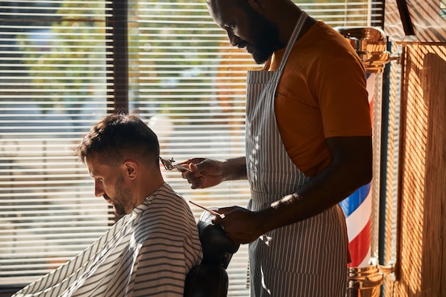 Bel barbiere maschio che taglia i capelli del cliente con un tagliacapelli in metallo