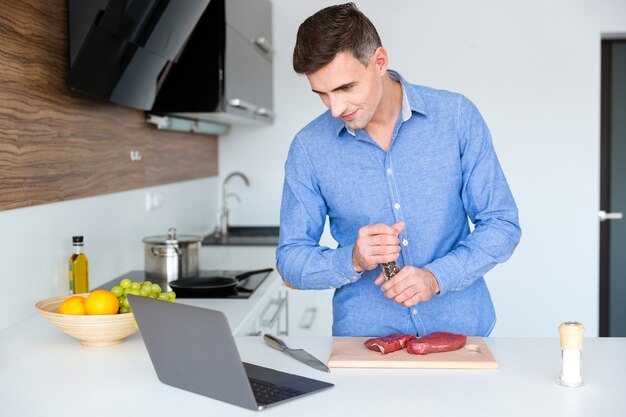 ラップトップで料理のレッスンを見て、キッチンで肉を準備する青いたわごとのハンサムな男性