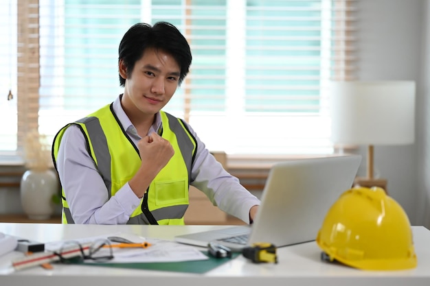 Handsome male construction engineer in yellow vest with arms raised celebrating success and smiling to camera