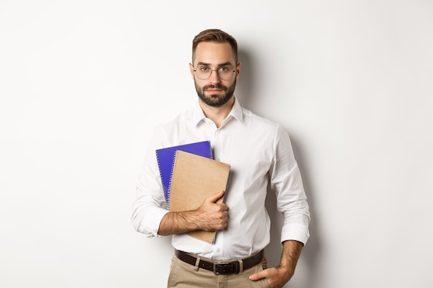 Handsome male employee holding notebooks, looking confident, standing over white background.