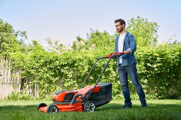 Un bel giardiniere maschio con i guanti che taglia il prato con un tosaerba elettrico in giardino durante il giorno