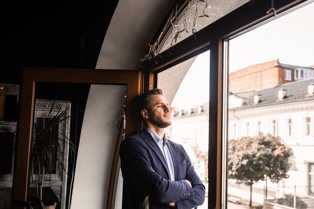 Handsome male model is posing in cafe. Bearded man weared suit stand and smile near window.