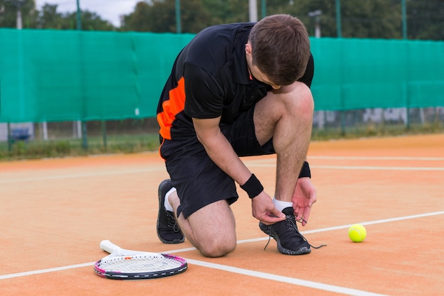 Photo handsome male tennis player tying shoelaces wearing a sportswear left racket and ball on a clay court outdoor in summer or spring