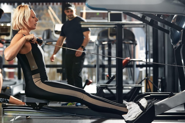 Handsome male trainer with athletic body helps girl client to perform proper exercises on the simulator The concept of training with a coach man