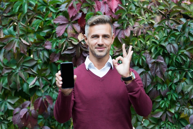 Handsome male using modern mobile phone. mature guy on autumn leaves background. fall business casual fashion. male show ok gesture. best quality ever. charismatic and successful man outdoor.