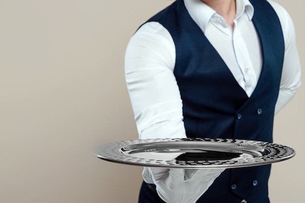 Photo handsome male waiter, white shirt, holds a silver tray, hand behind his back. concept of wait staff serving customers in a restaurant.