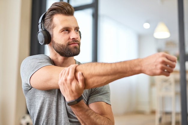 Handsome male in wireless headphones looking away and smiling while doing exercise at home