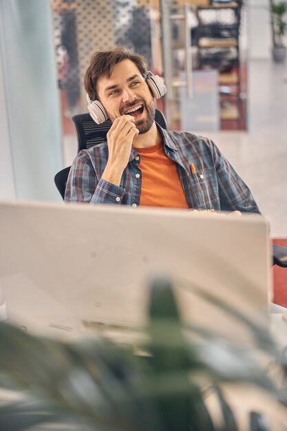 Handsome male worker in headphones enjoying food and smiling while sitting at the table with computer in office