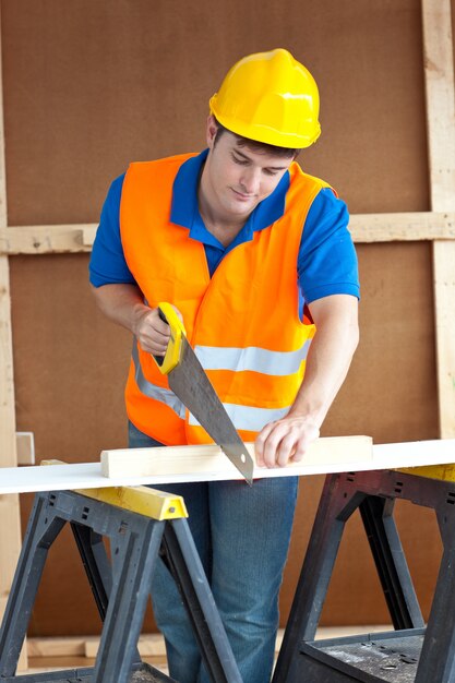 Handsome male worker wearing a yellow hardhat sawing a wooden board