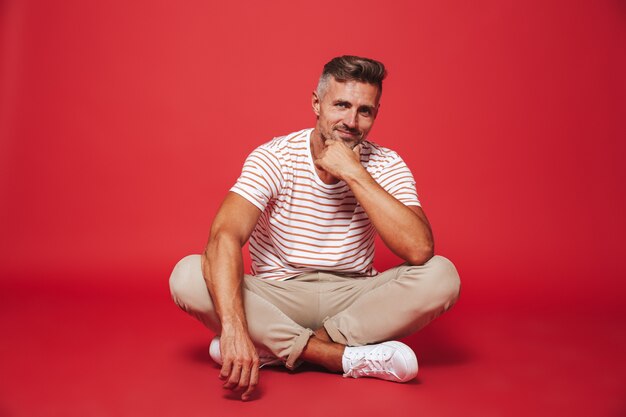 handsome man 30s in striped t-shirt smiling, while sitting on floor with legs crossed isolated on red