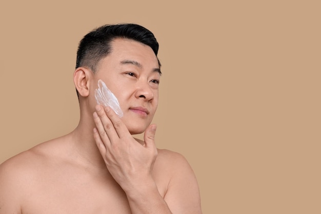 Handsome man applying cream onto his face on light brown background Space for text