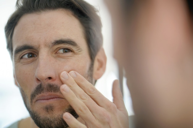 Photo handsome man applying face cream looking in mirror