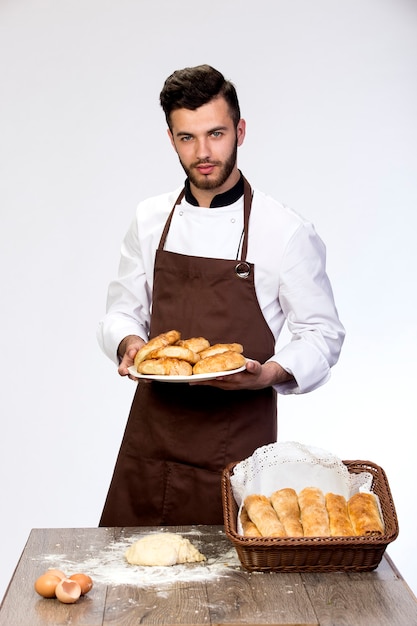 Uomo bello in grembiule con prodotti da forno freschi isolato sul muro bianco.