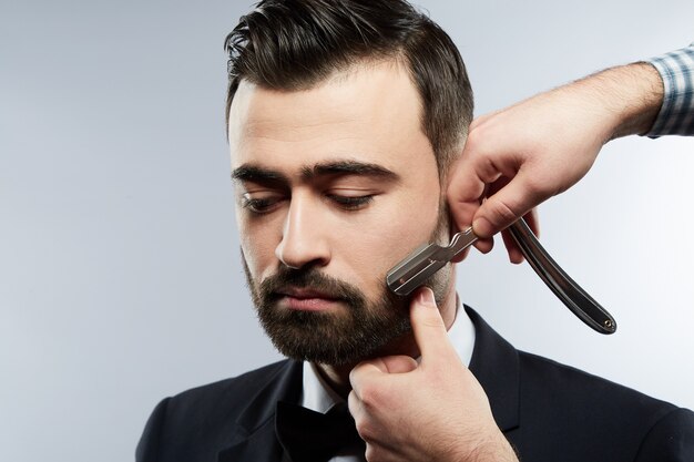 Handsome man at barbershop wearing blak suit and tie, man's hands wearing shirt making a beard form  for man with black hair at studio background.