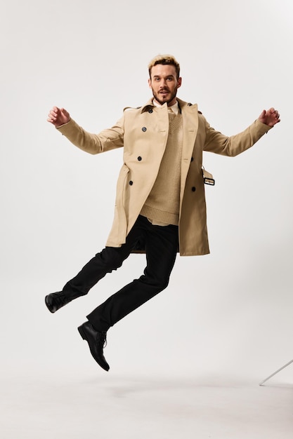 Красивый мужчина в бежевом пальто в осеннем стиле прыгает в студии на светлом фоне Фото высокого качества