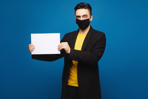 Uomo bello in una maschera protettiva nera che tiene un foglio di carta vuoto e che esamina la macchina fotografica, isolata ai precedenti blu. concetto di promozione. concetto di assistenza sanitaria