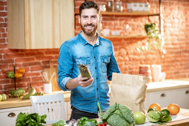 Bell'uomo in camicia blu che disimballa cibo sano dalla borsa della spesa in cucina