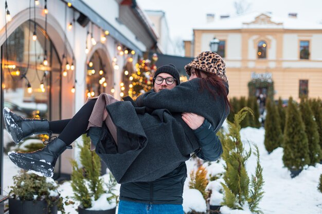 Handsome man carries his girlfriend in his arms in the city on a winter day Happy couple of lovers in fashion winter clothes having fun together