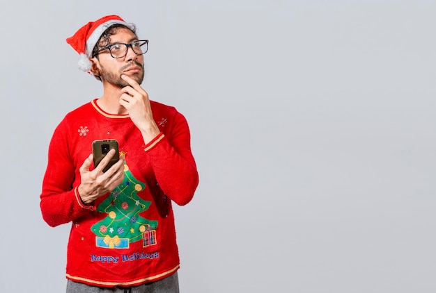 Handsome man in christmas clothes thoughtful holding cell phone Thoughtful man with christmas hat holding cell phone Young man in christmas clothes thoughtful with cell phone isolated