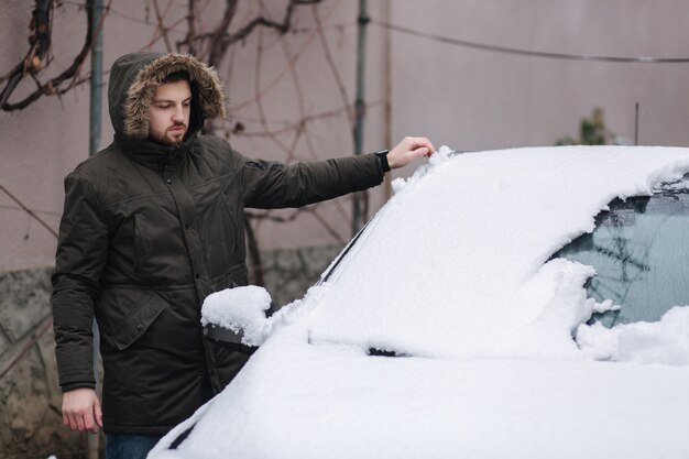 ハンサムな男は手を使って雪から彼の車をきれいにします。冬の屋外