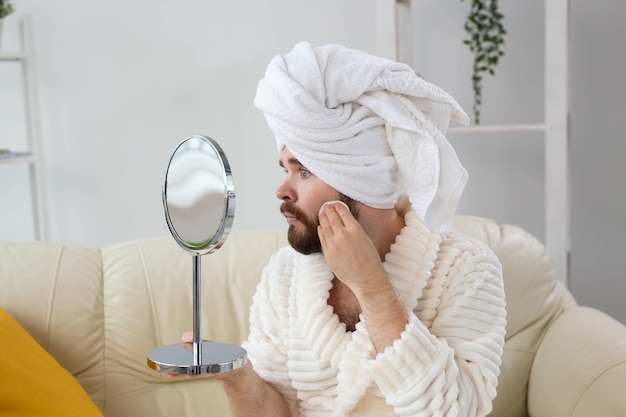 Handsome man cleaning face skin with batting cotton pads and\
looking at mirror. spa, body and skin care for male concept.