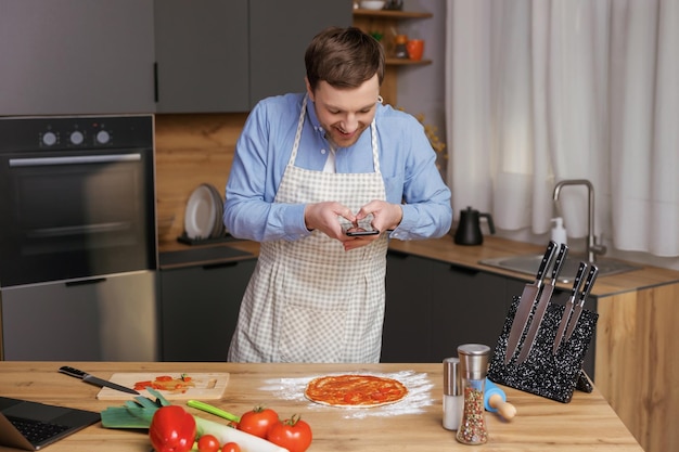 キッチンで料理をし、携帯電話で食べ物を準備する写真を作るハンサムな男性