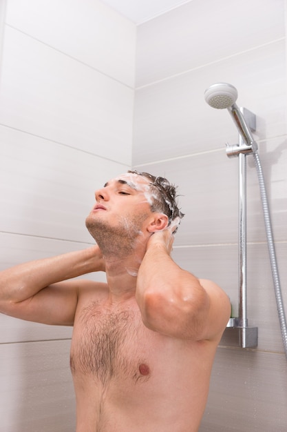 Handsome man diligently washing his dripping foam hair in shower cabin in the modern tiled bathroom