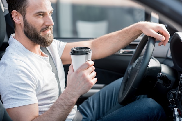 行くためにコーヒーで車を運転する白いTシャツを着たハンサムな男