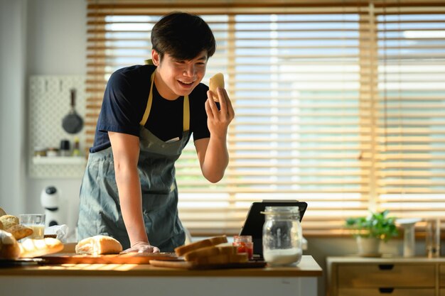 Handsome man dressed in casual clothes holding fresh bread standing at dining table in kitchen and having video call over laptop