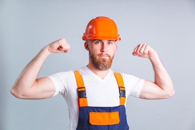 Handsome man engineer in building protective helmet on gray wall looks to the front and shows muscle biceps