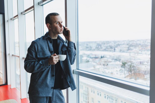 A handsome man of European appearance in a casual suit drinks coffee talks on the phone and looks out the window