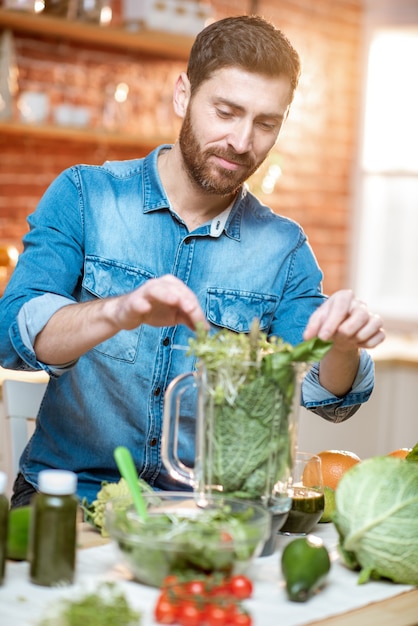 ハンサムな男がブレンダーに緑のローフードを入れ、自宅のキッチンで野菜のスムージーを作る