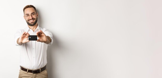 Handsome man in glasses holding a credit card smiling pleased standing over white background person