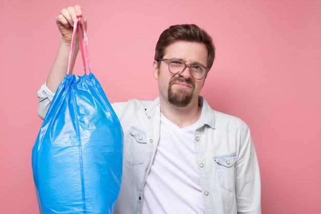 Красивый мужчина держит синий мешок для мусора на розовом фоне
