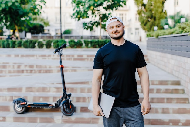 Handsome man holding laptop in his hand standing near electric scooter Active lifestyle backpacker traveler