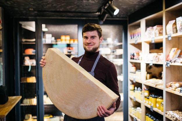 A handsome man holds a large piece of Maasdam cheese in his hand Cheese with big holes Cheese shelf background