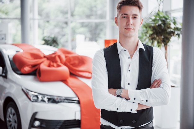 A handsome man is a buyer standing next to a new car at the dealer center and looking at the camera.