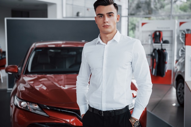 A handsome man is a buyer standing next to a new car at the dealer center and looking at the camera.