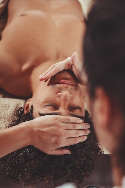 Photo handsome man is lying down on massage table and getting healthy facial massage by young female therapist in the spa center.