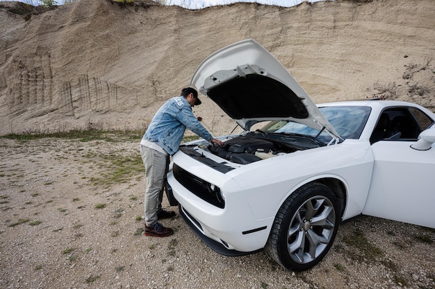 Handsome man in jeans jacket and cap is standing near his white muscle car with opening hood check oil level of engine