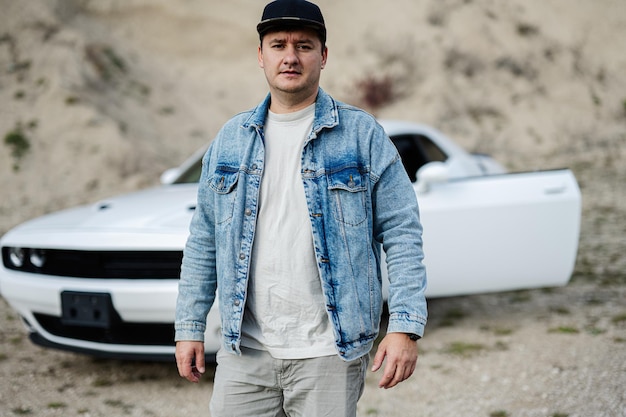 Handsome man in jeans jacket and cap walking near his white muscle car in career