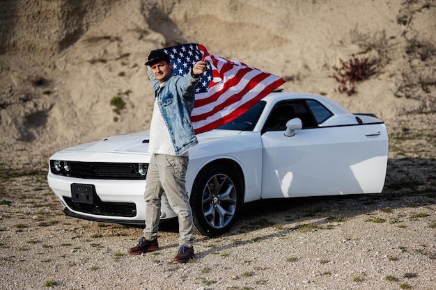 Красивый мужчина в джинсовой куртке и кепке с флагом США рядом со своим белым американским маслкаром в карьере
