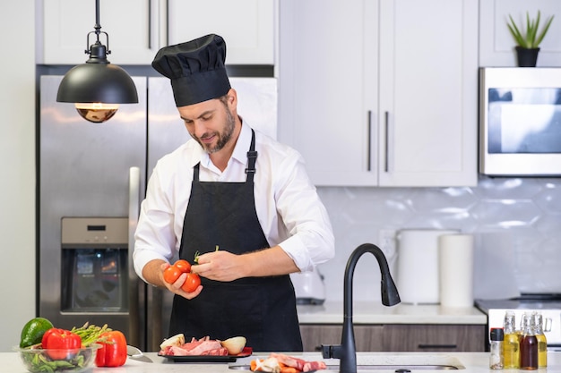 Handsome man in kitchen is preparing healthy fruits salad and meat man in apron and chef hat prepari