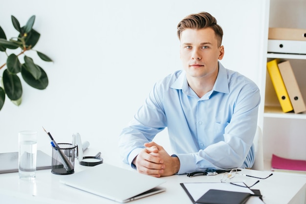 Handsome man looking at camera while sitting with clenched hands near laptop