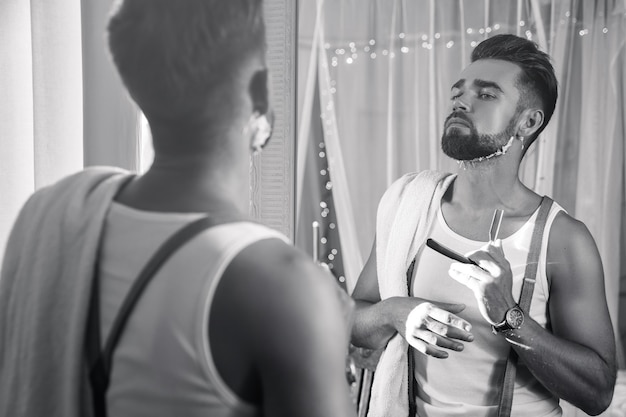 Handsome man looking in the mirror and shaving his beard with a straight razor