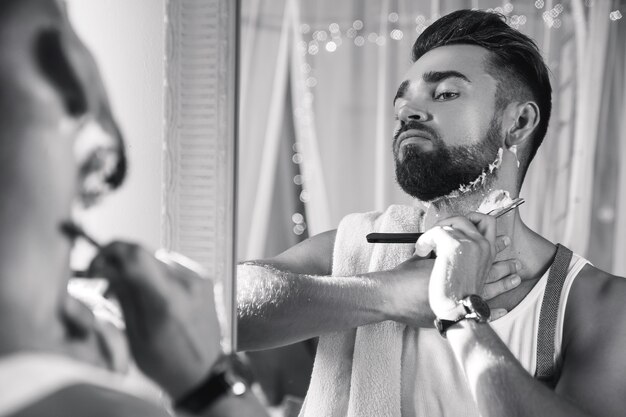 Handsome man looking in the mirror and shaving his beard with a straight razor