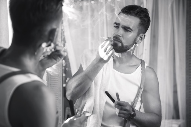 Handsome man looking in the mirror, smoking a cigarette and shaving his beard with a straight razor